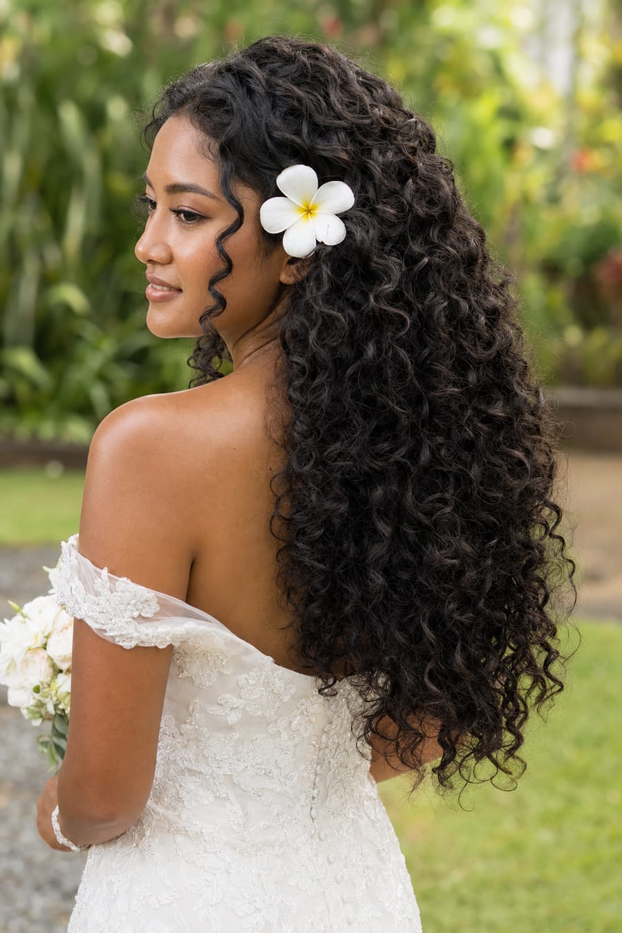 Indonesian bride with long natural 3a curly hair worn loose, a white frangipani above her ear, three-quarter back view
